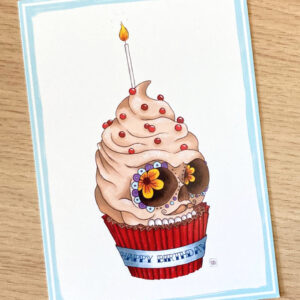 carte postale représentant un cupcake en forme de crâne mexicain avec une bougie et un "happy birthday" écrit sur le papier du cupcake