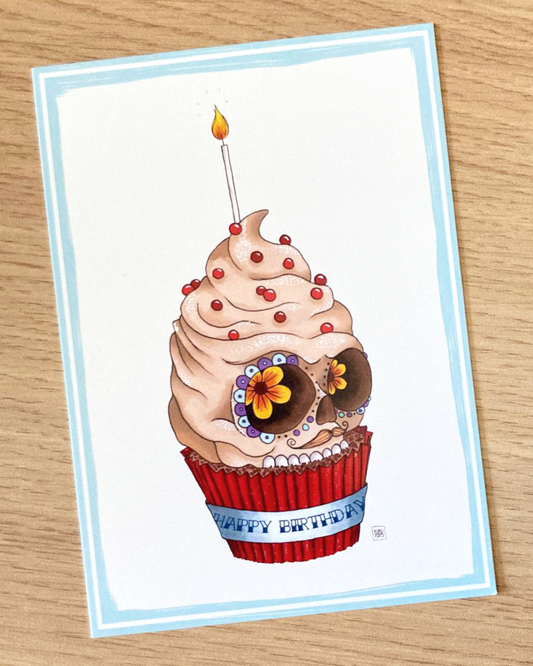 carte postale représentant un cupcake en forme de crâne mexicain avec une bougie et un "happy birthday" écrit sur le papier du cupcake